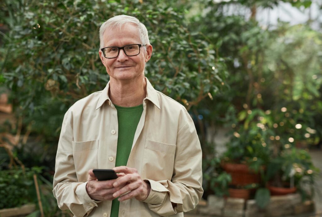 Senior man standing in a greenhouse garden, smiling while using a smartphone. Natural sunlight.